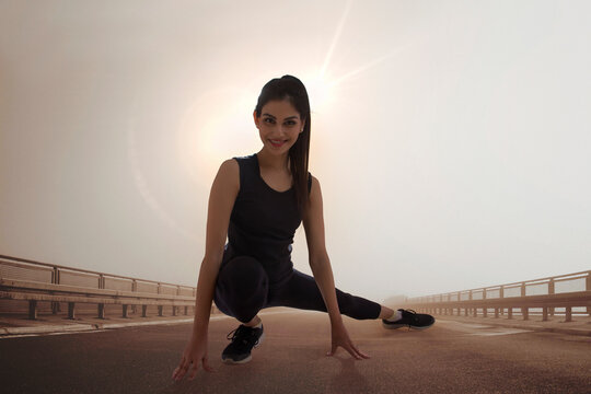 Young woman in workout clothes doing stretching exercises outdoors with sun in the background.