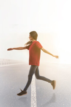 Young woman leaping joyfully in mid air across the road with sun in the background.