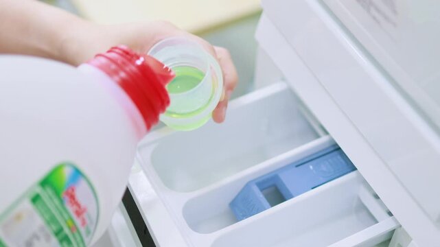 A close-up of a person's hand pouring liquid laundry detergent into the detergent drawer of a white washing machine.