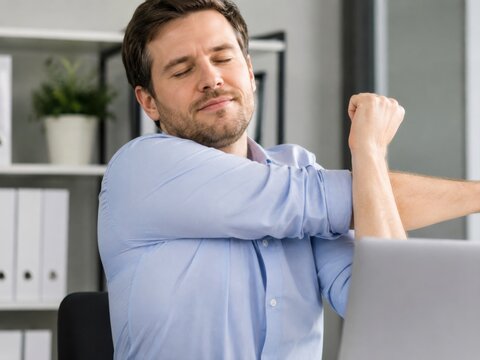 A man in a light blue shirt stretching his arms in an office, eyes closed in relief while sitting.
