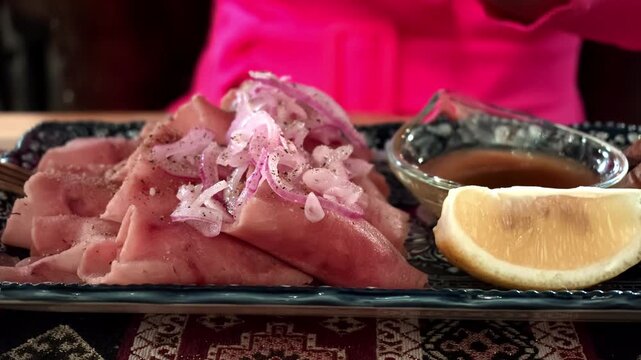 Closeup view of thin sliced ham layered with onion rings on decorative plate, paired with dipping sauce and lemon wedge, against soft pink blurred background for casual dining scene