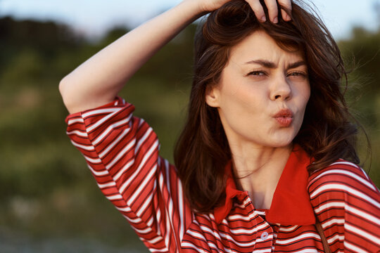 Young brunette woman in a red striped shirt poses playfully outdoors, pouting her lips towards the camera during a golden hour sunset to capture a fun and authentic candid moment.