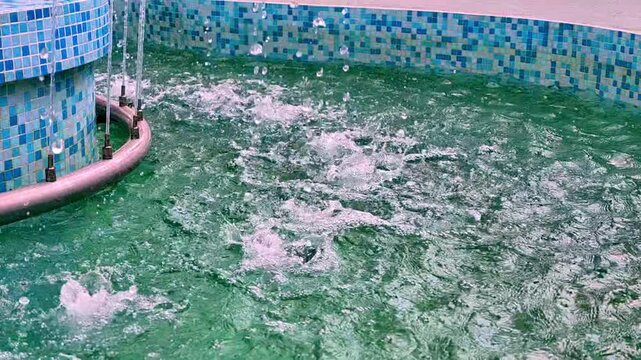 Indoor pool interior with curved tiled edge, blue mosaic walls, and turquoise fountain jets bursting upward, creating droplets, ripples, spray, reflections, and shimmering color, with indoor, pool