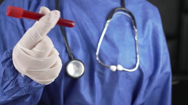 Gloved medical professional in blue scrubs holds a blood sample tube close to the camera.