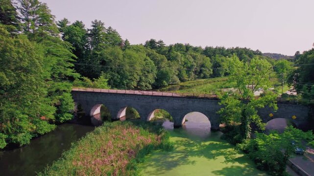 Drone flies back to reveal wide view of old stone arch rail road bridge on the Assabet River reservoir, in Northborough, Massachusetts.