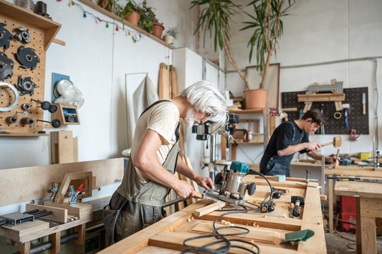 Concentrated master technician setting up handheld wood router instrument for work in workshop, professional carpenter restorer preparing electric plunge router machine at workbench in studio.