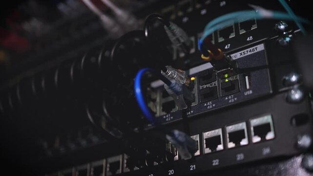 A close-up of network cables and ethernet ports on a server rack in a dark data center with blue glowing lights.