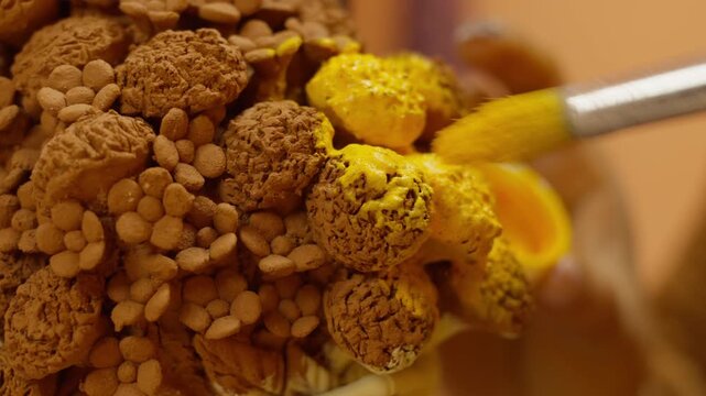 Closeup of artisan painting clay marigold bouquet on Catrina sculpture with yellow paint in small dish