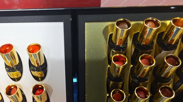 Display of multiple lipsticks with golden cases and varying red to nude shades, arranged for retail viewing in a cosmetic store setting, presenting diverse makeup product choices.