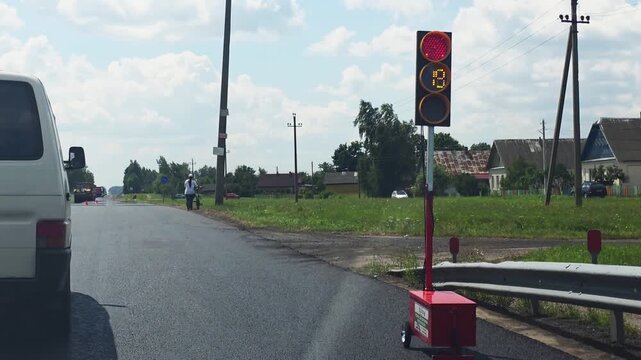 Portable traffic light displaying red signal with countdown during road repair on rural highway, white van stopped, worker visible in distance, sunny day, clear sky, reverse movement