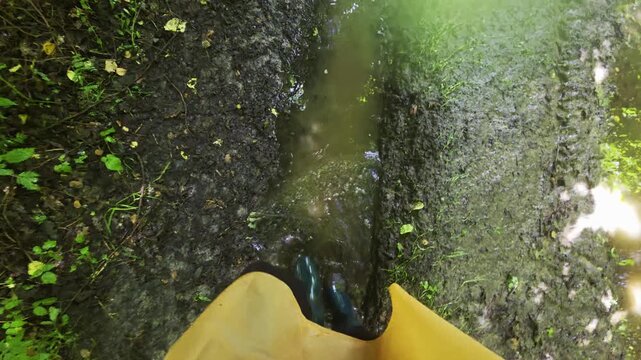Person point of view of boot stepping in mud and puddle on wet path during daytime walk outdoors, individual wearing yellow raincoat and green rubber boot, nature environment, slow motion