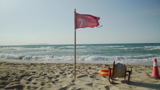 A red warning flag flutters in the strong wind on a sandy beach, signaling danger due to rough sea waves and currents.