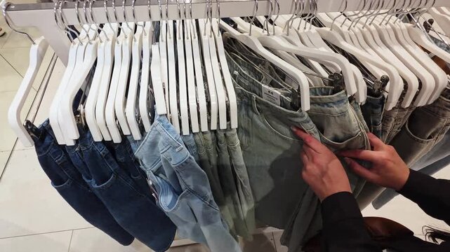 Person hands browsing through collection of denim jeans on hangers in clothing store, selecting item for purchase, fashion retail shopping experience