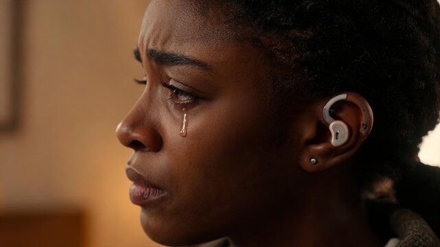 A close-up portrait of an emotional adult Black woman crying, wearing a hearing device in an indoor space.