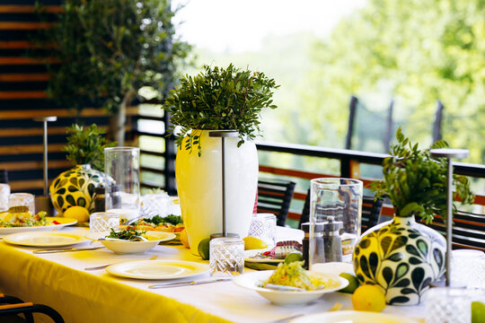 Stylish dining table setup on a terrace, featuring fresh food, dinnerware, and green plants, ready for an outdoor lunch