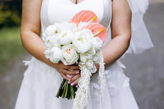 Bride standing in a white dress, holding a beautiful floral wedding bouquet featuring white peonies, roses, and coral anthurium