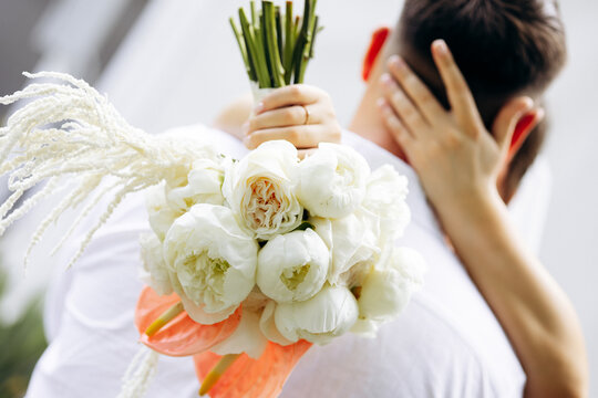 Celebrating marriage, a bride embraces her groom, holding a beautiful white rose and peony wedding bouquet with anthurium