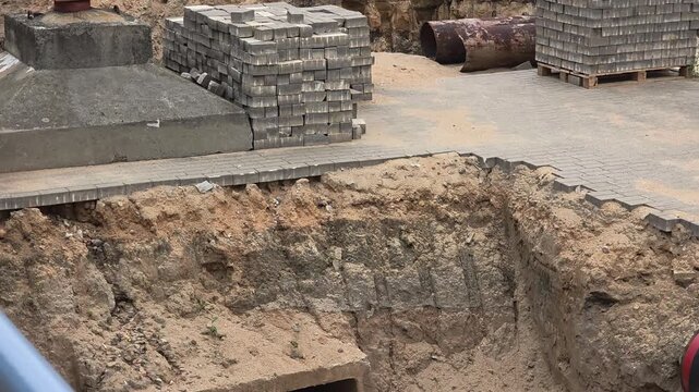 Construction site with deep trench excavation for underground pipe installation and new paving stones laid on surface during infrastructure development work.