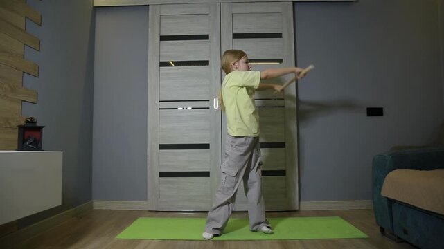 Caucasian child performing physical exercise with wooden stick in living room of modern house, engaging in workout routine for health and fitness practice indoors