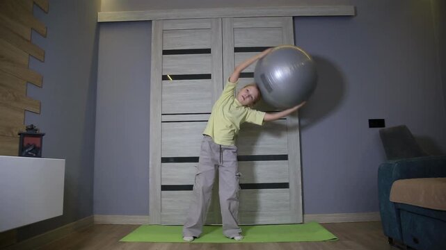 Young girl performing fitness exercises with stability ball on yoga mat indoors. Child engages in physical activity, body movement, healthy lifestyle for well-being.
