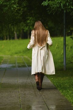 Woman with long hair walks away down stone path in green park. Grass and trees line the way. Water glistens on the ground from recent rain