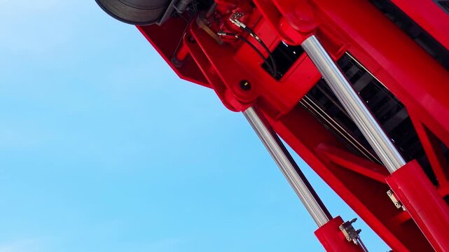 Red hydraulic machinery components against clear blue sky, featuring shiny silver hydraulic cylinders and black hoses, indicating heavy equipment and industrial power