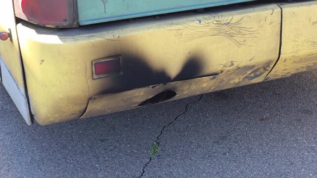 Rear view of old worn vehicle bumper showing extensive damage, scratched yellow paint, dents, and prominent black soot stain around exhaust pipe exit on asphalt ground