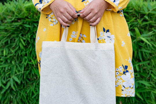 Woman Holding Blank Cotton Tote Bag in Yellow Floral Dress Against Green Foliage