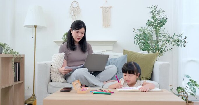 Asian single mother checking worksheet beside laptop while preschool daughter writes in workbook at table, coordinated home study routine, parent support, early education, child growth, family care