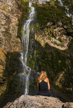 Woman sits on a rock looking at a beautiful waterfall with a small rainbow on the mossy cliff during a summer hike