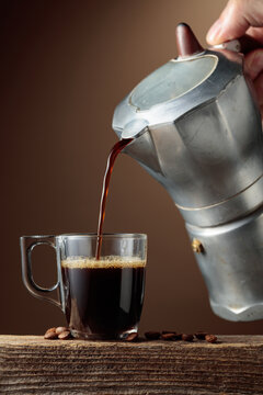 Black coffee is poured from a classic Italian aluminium moka pot into a glass coffee cup.