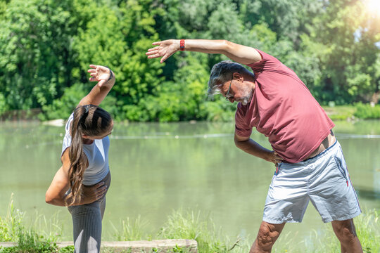 Stretching senior man and fitness coach performing a standing side stretch beside a lake to improve flexibility, spinal mobility 
