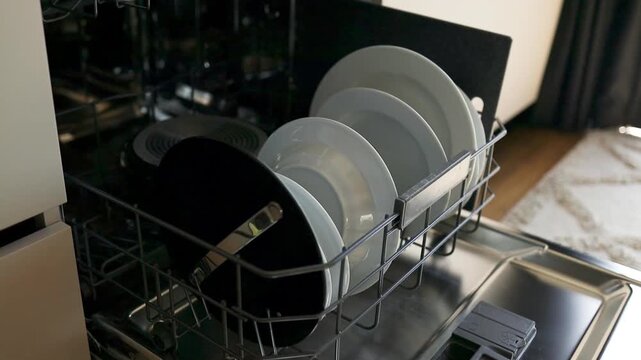 An open automatic dishwasher machine packed with clean white plates and dishes after a washing cycle. The shot captures a modern kitchen interior, efficient housekeeping, routine home chores, and