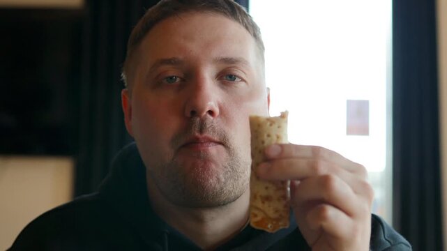 A young caucasian man with a beard is eating a fresh homemade pancake stuffed with apple filling. He looks thoughtful or relaxed while enjoying a sweet crepe or pastry for breakfast or dessert in a