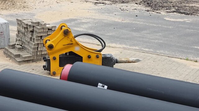 Heavy duty yellow hydraulic breaker attachment for excavation machinery rests on paved ground at construction site next to large black pipes and stack of grey paving stones