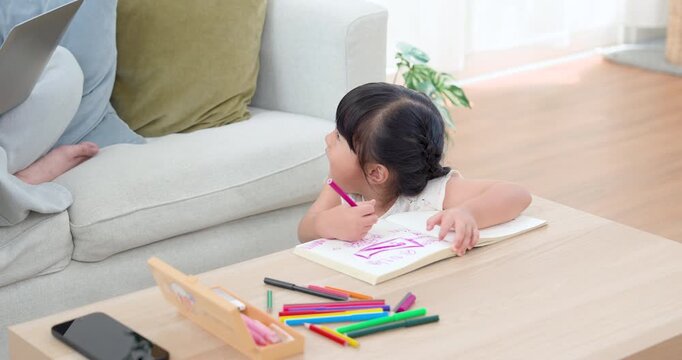 Preschool Asian daughter pauses drawing and looks toward single mother, holding pink pencil above workbook on table, expressing trust, encouragement, connection, and growing home learning progress.