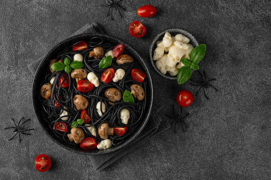 Halloween-themed food. Black squid ink spaghetti with mozzarella skulls, mushrooms, cherry tomatoes and fresh basil in a bowl. Creative spooky food styling for festive autumn celebrations. Top view.