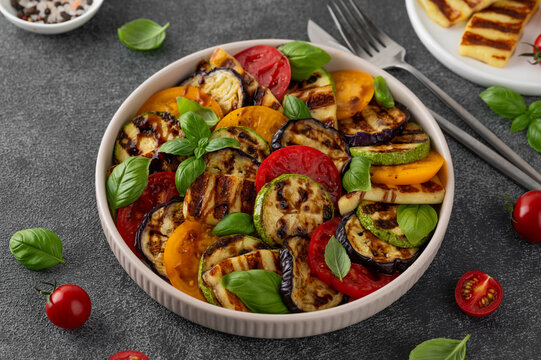 Grilled vegetable salad with zucchini, eggplant, tomatoes, halloumi cheese, basil and balsamic glaze on a plate on a dark concrete background. Healthy food.