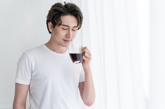 Portrait of young handsome asian man hands holding coffee glass cup morning spring time in white bedroom. Happy cheerful relaxing in summer. Korean male guy wakeup, university asia lifestyle concept