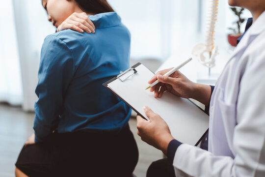 Woman suffering from back and shoulder pain during a medical consultation with a doctor writing notes on a clipboard, seeking diagnosis and treatment for her musculoskeletal discomfort