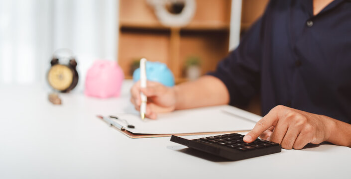 Person taking notes and calculating expenses on a desk with a calculator, managing finances, budgeting, and planning future savings with piggy banks in the background