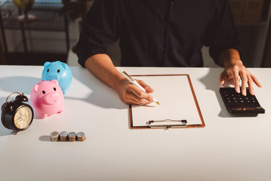 Person calculating finances and managing budget, planning future savings with piggy banks, coins, and an alarm clock on a white desktop