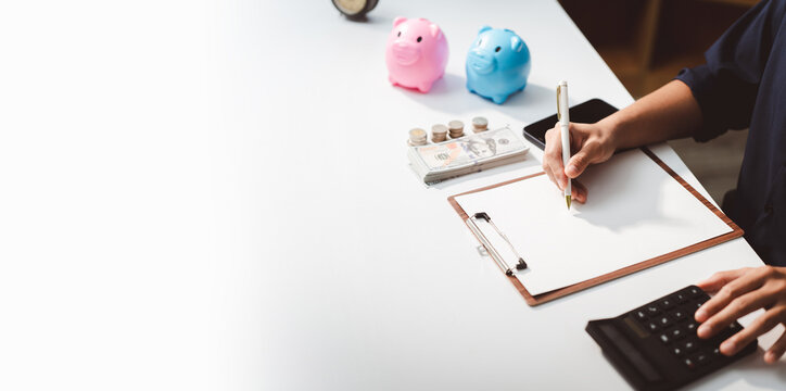 Person calculating expenses, planning budgets, and managing finances with money, coins, and piggy banks on a white desk, representing savings and investment strategies