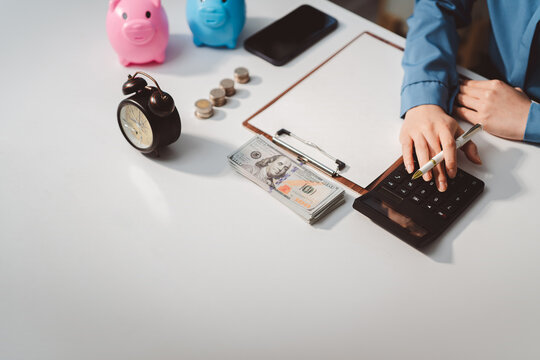 Person calculating finances on a desk with money, piggy banks, and an alarm clock, symbolizing saving time and financial planning for the future