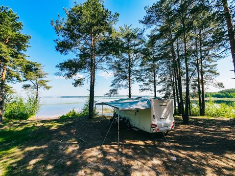 White camper trailer positioned under tall pine trees near tranquil lake on clear sunny day