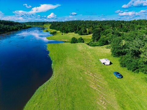 Aerial view of a camper parked beside a river surrounded by lush green landscapes in summer