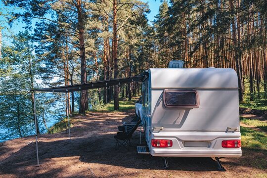 RV parked under tall trees by the lake with an extended awning and camping setup