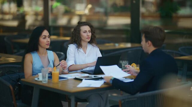 Three diverse professionals are engaged in an outdoor business meeting at a cafe. The team discusses important documents and works together in a collaborative environment.