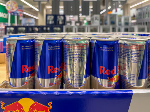 Berlin, Germany July 29, 2026. Rows of Red Bull energy drink cans fill a case with blurred store shelves behind. The silver and blue cans display nutrition information beneath the bright red logo.