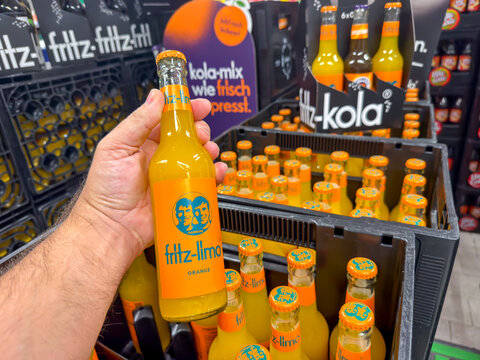 Berlin, Germany July 29, 2026. A hand holds an orange labeled fritz limo soda bottle in a grocery store aisle. Crates of fritz kola and more fritz limo bottles line the shelves in the background.